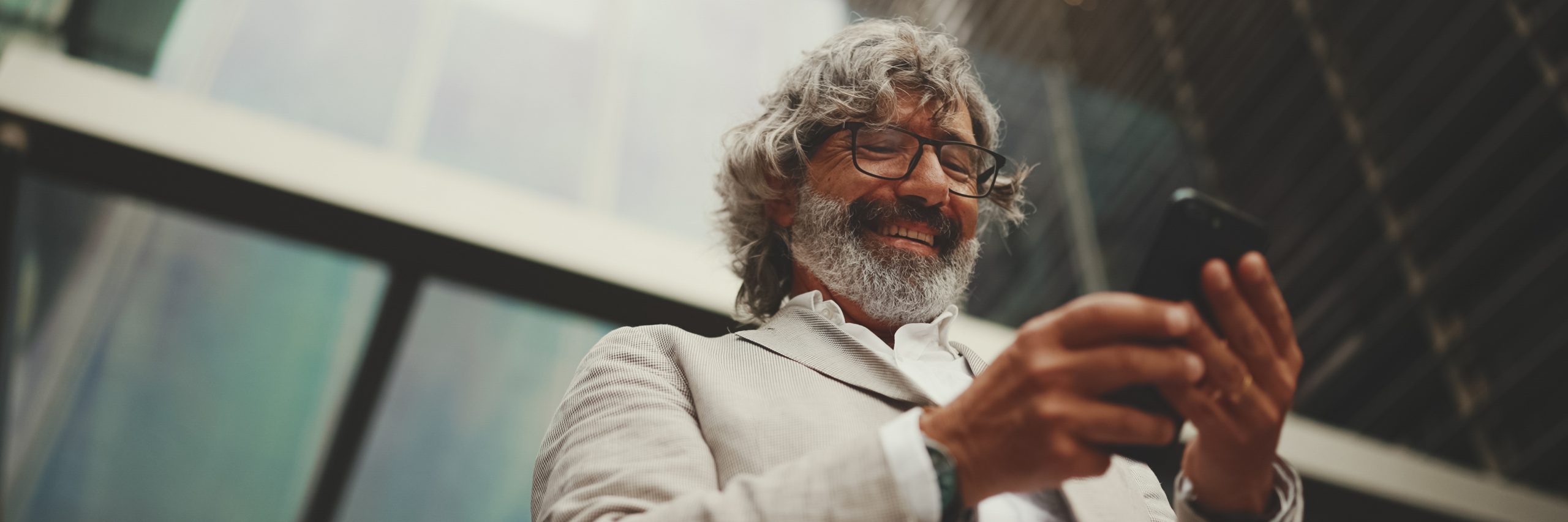 Mature businessman with beard in glasses stands on the street at the business center background, Panorama. Middle aged manager texting message scrolling tapping to smartphone. Joy from good deal